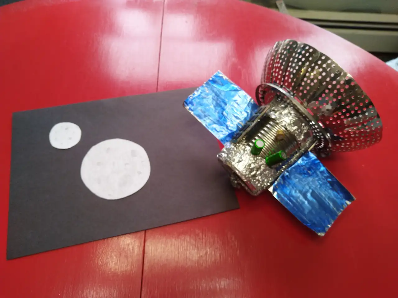 A simple sculpture resembling a satellite, constructed from a metal can, aluminum foil, a vegetable steamer basket, and miscellaneous electronic components. Next to the sculpture are two circles of white paper on a black sheet of paper, resembling two planets or moons in space.