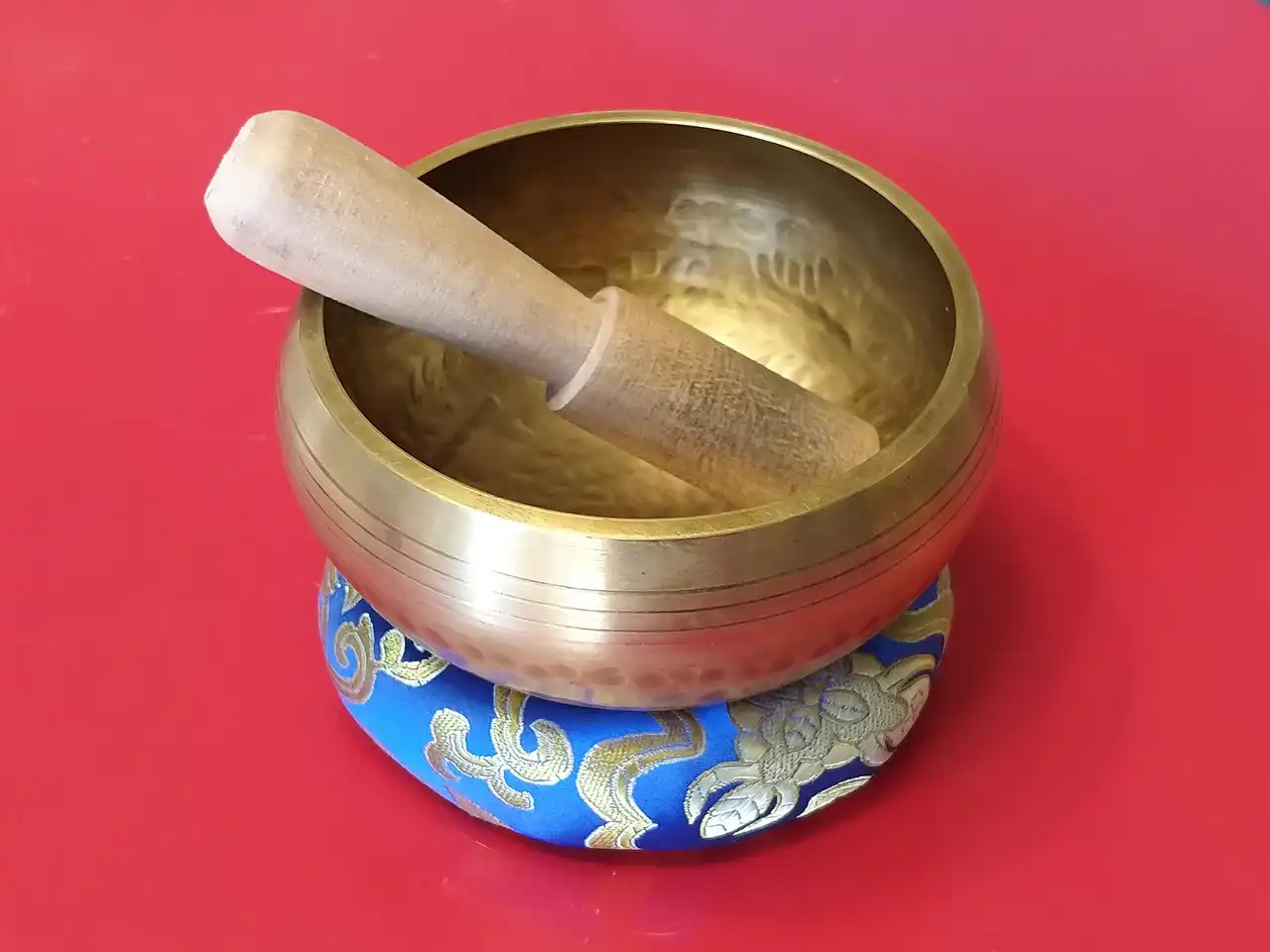 A Tibetan singing bowl: a brass-colored concave metal bowl resting on a small blue cushion. The bowl contains a wooden mallet.